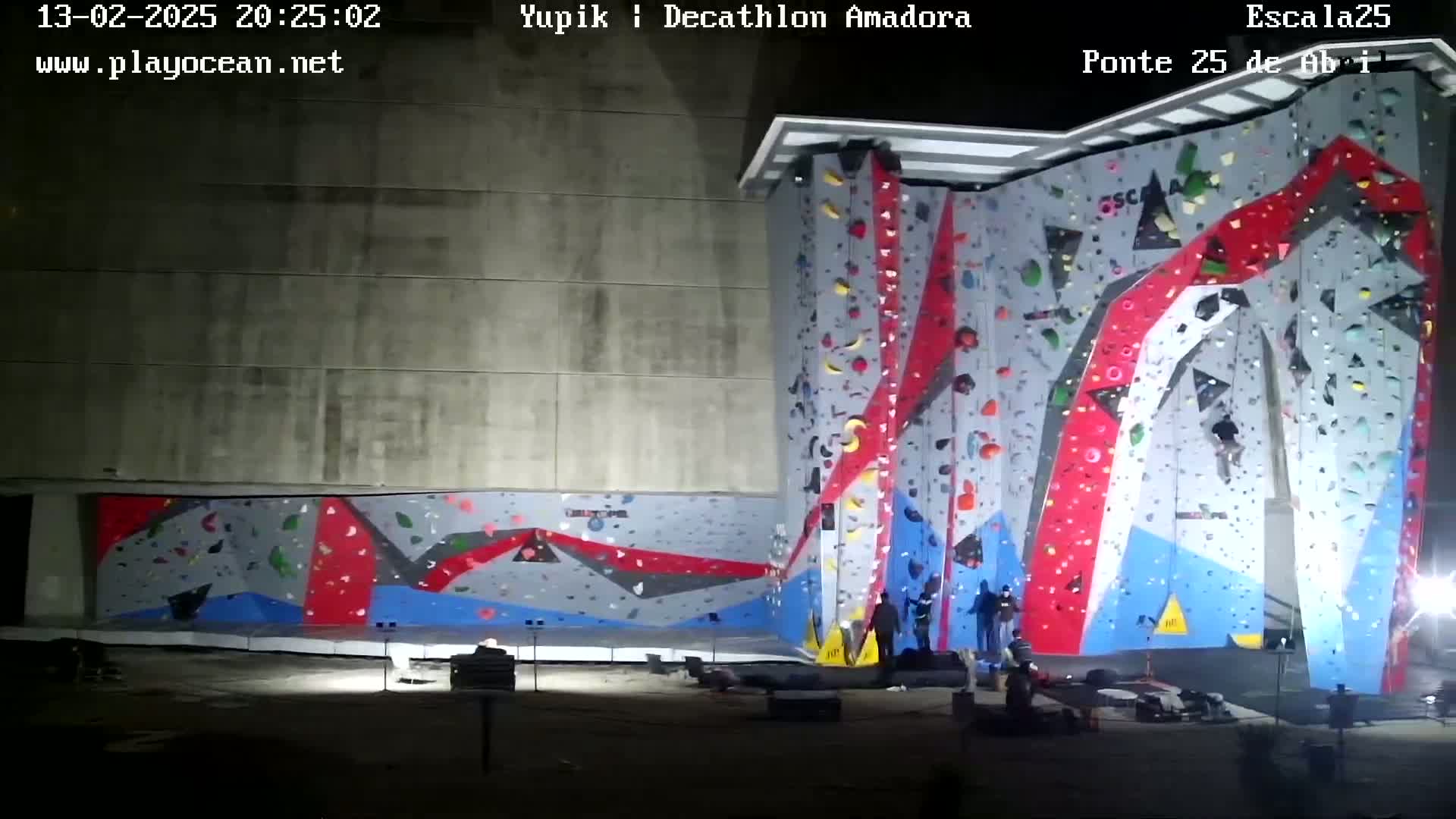 At night, several people are gathered around a large, brightly colored outdoor climbing wall.