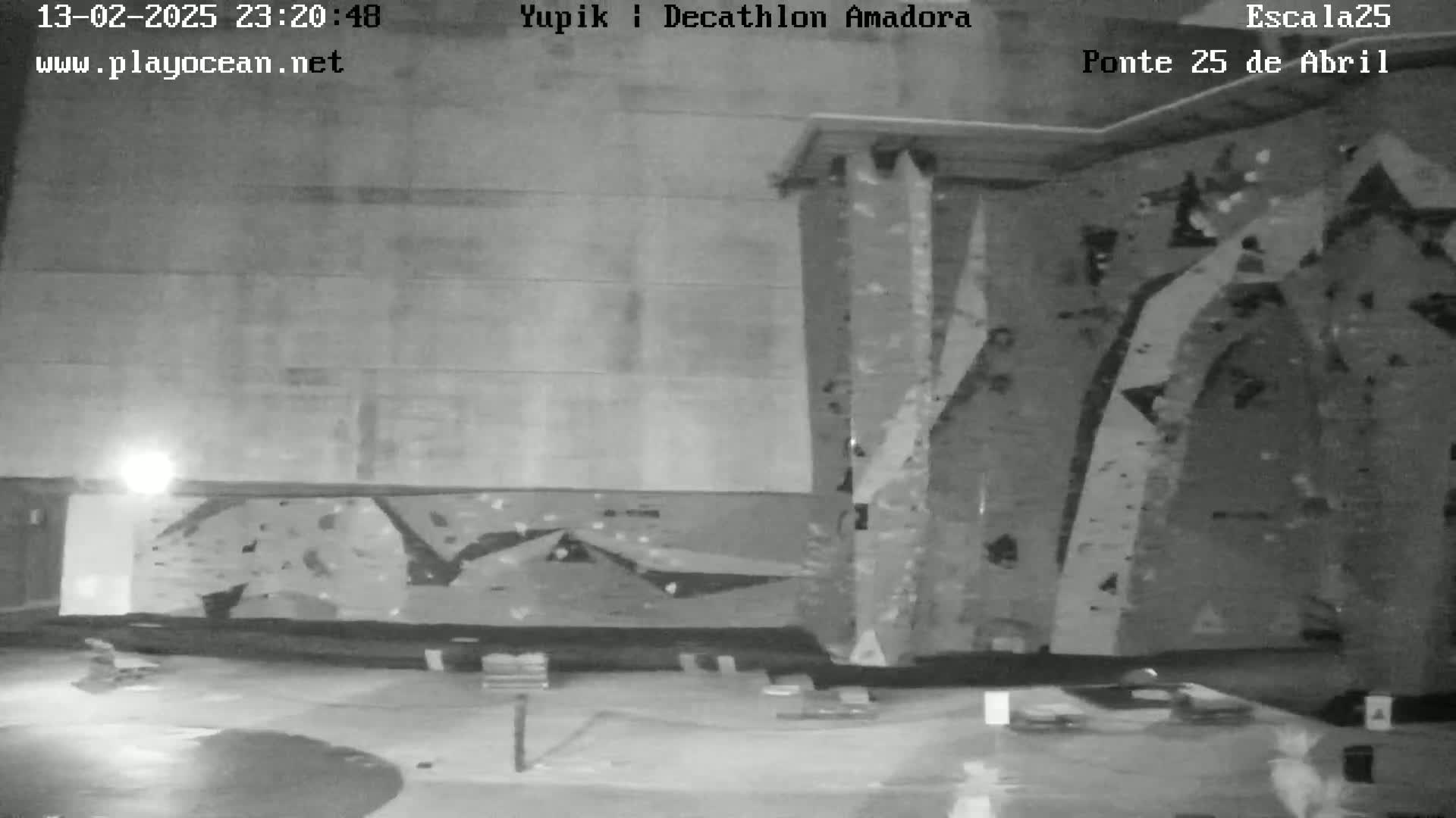 A nighttime, black and white image shows an indoor climbing wall with various holds and a mostly empty floor.