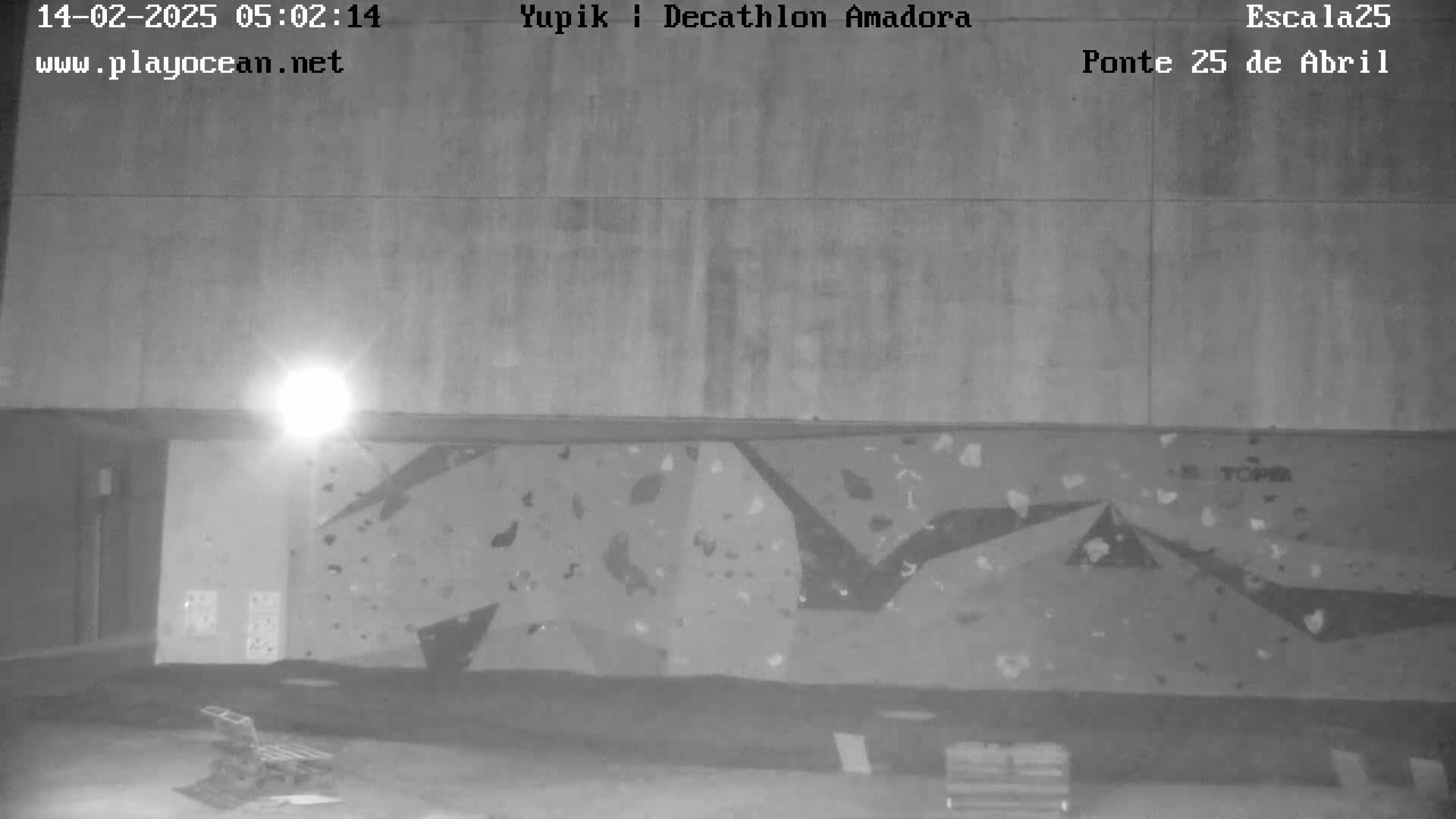 A nighttime, low-light image shows an indoor climbing wall with various holds, lit by a single overhead light.