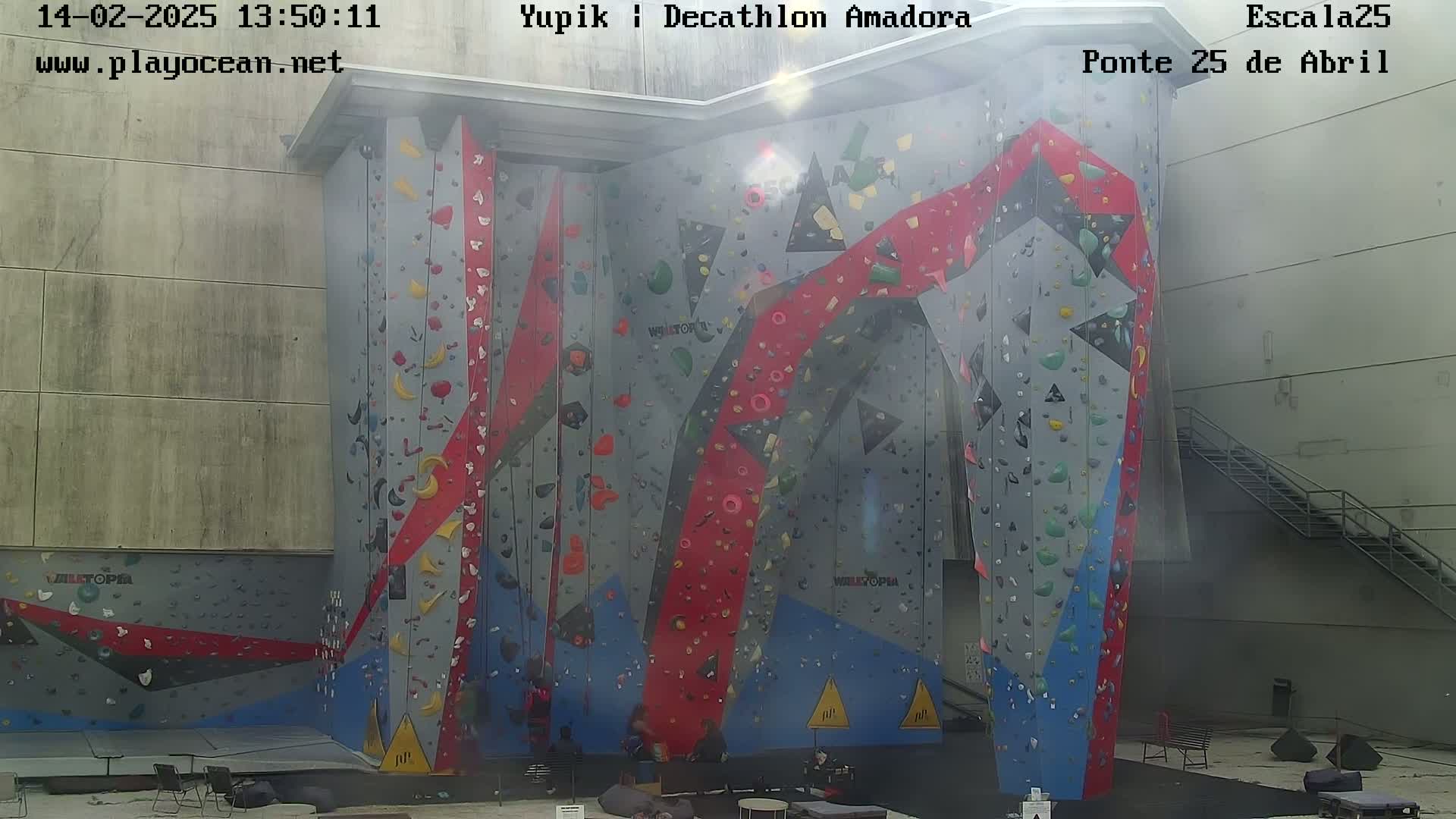 A large indoor climbing wall with various colored holds and geometric designs is shown in hazy sunlight.