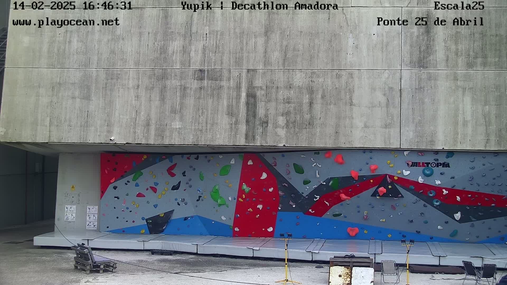 An outdoor climbing wall with various colored holds is situated against a large concrete wall.