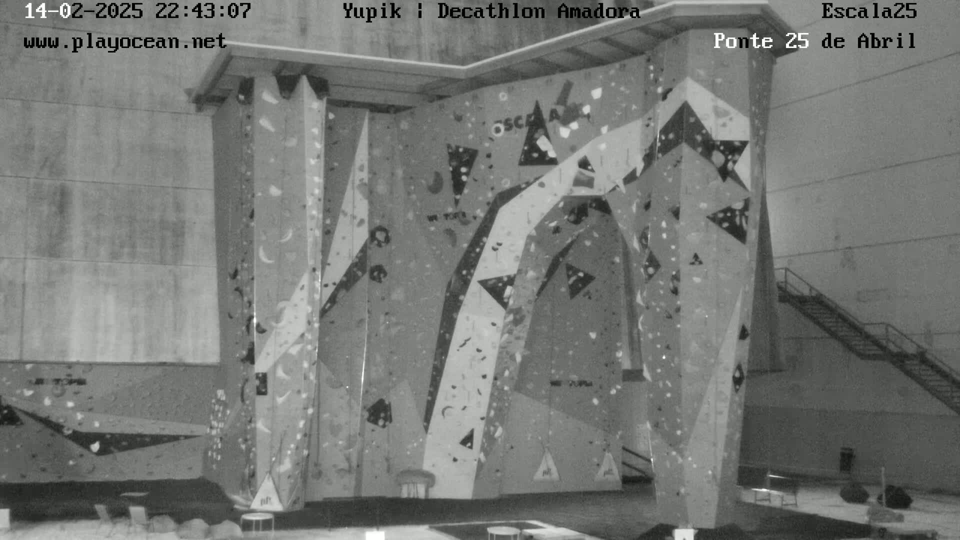 A grayscale image shows an indoor climbing wall with various holds and routes, under artificial lighting.