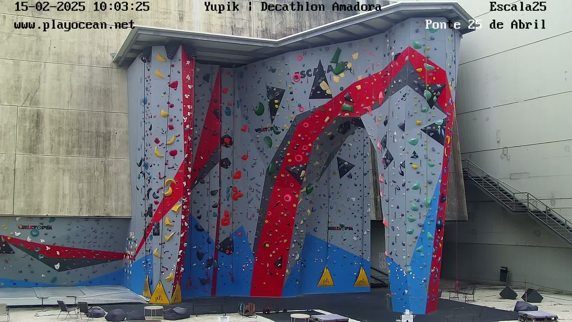 A large, colorful indoor climbing wall with various holds and a gray concrete building in the background.
