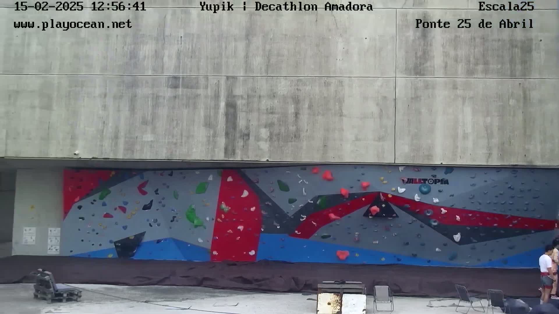 A large, colorful outdoor climbing wall is situated against a gray concrete building, with a few people standing near it.