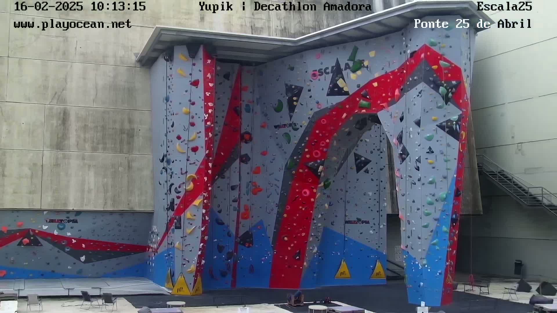 A large, multi-colored indoor climbing wall with various holds is situated against a light gray building.