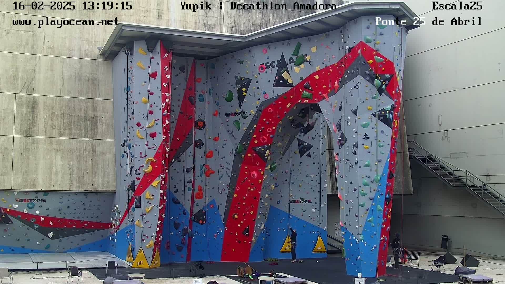 A large, multi-colored indoor climbing wall with a few people near the base, under an overcast sky.