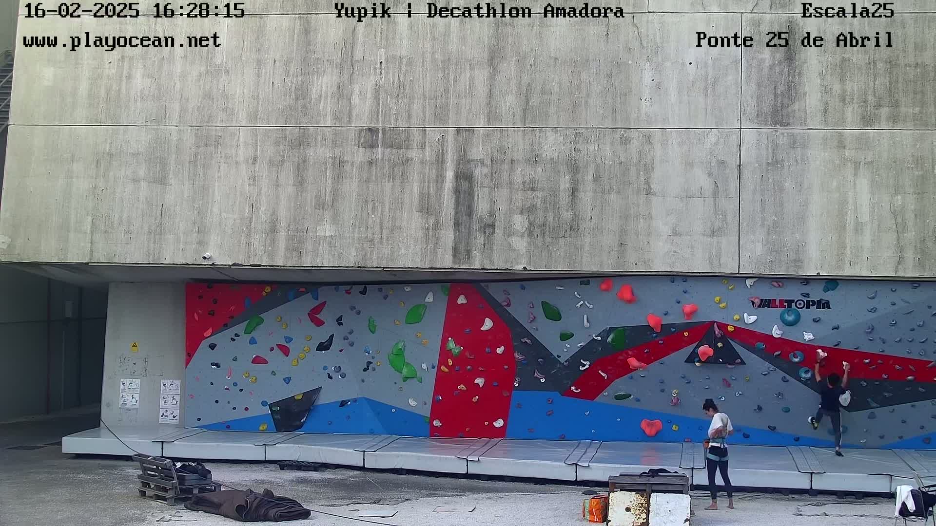 An outdoor climbing wall with two people climbing and a gray concrete building behind it.