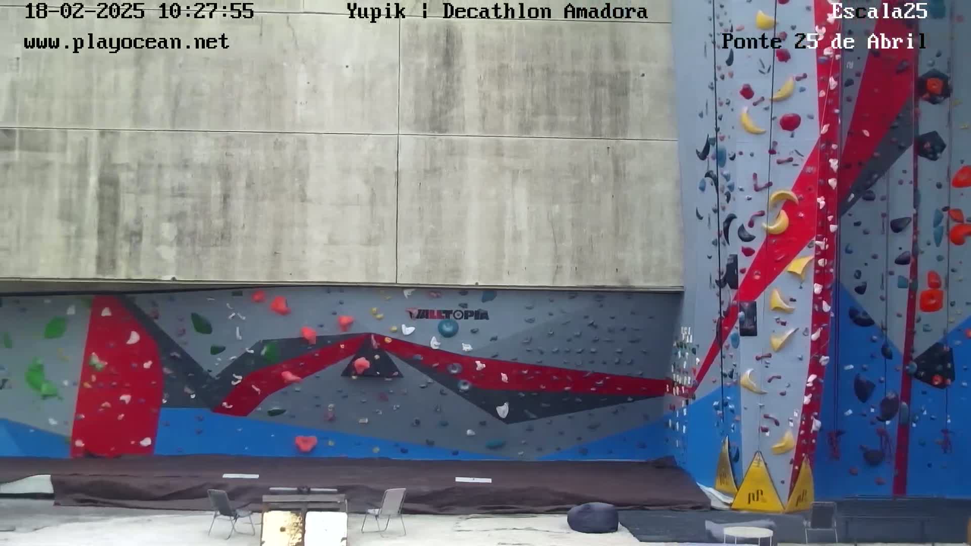 An outdoor view of two large climbing walls with various colored holds, situated against a concrete building on a mostly sunny day.