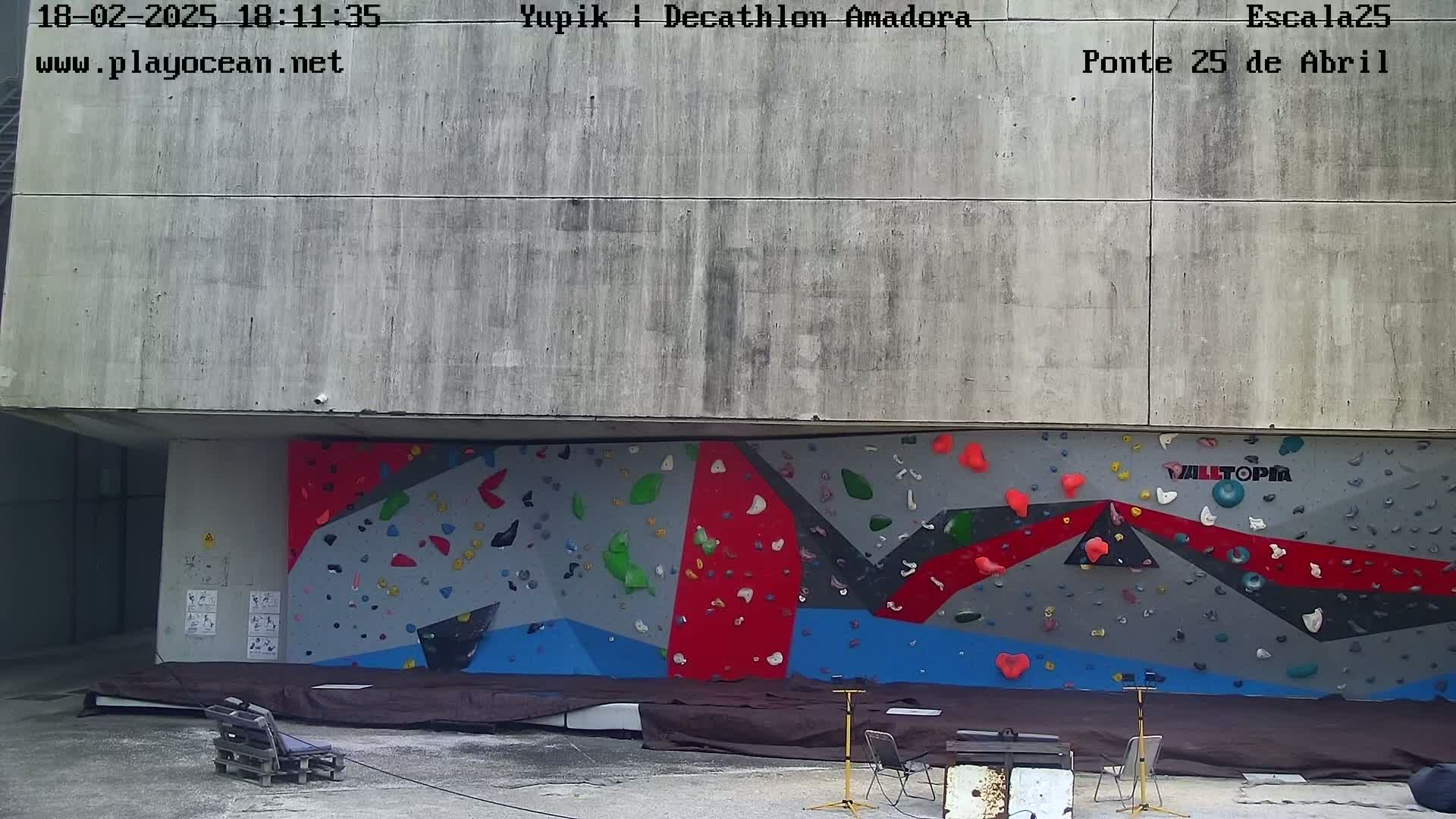 An outdoor climbing wall with various colored holds is situated beneath a concrete overhang.