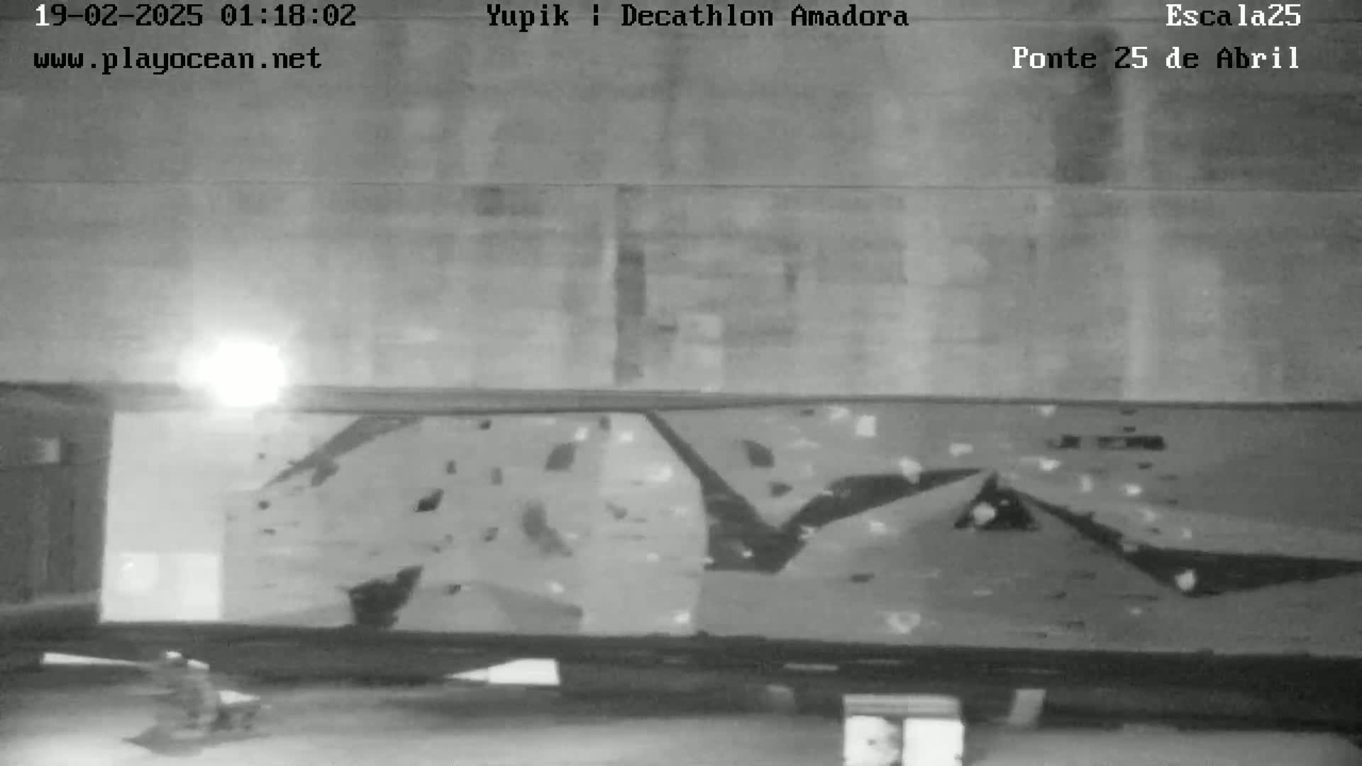 A nighttime, low-light grayscale image shows a person standing near a large climbing wall indoors.
