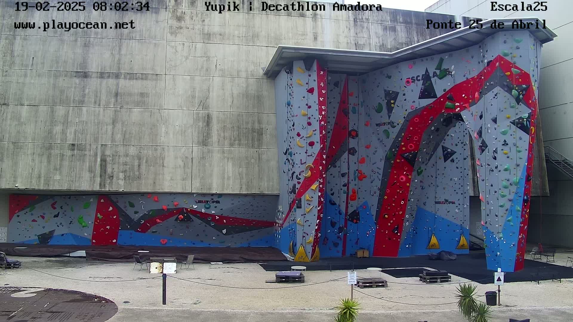 An outdoor climbing wall with red, blue, and gray holds is situated against a concrete building on a partly cloudy day.