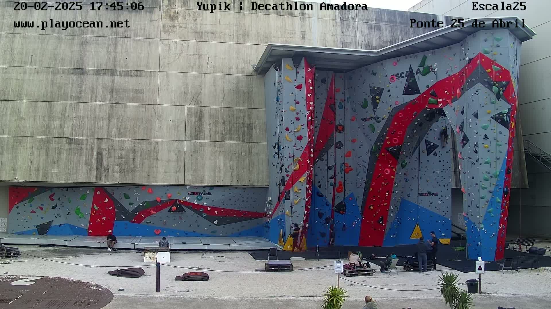 An outdoor climbing wall with various colored holds is situated next to a large gray building on a mostly clear day.