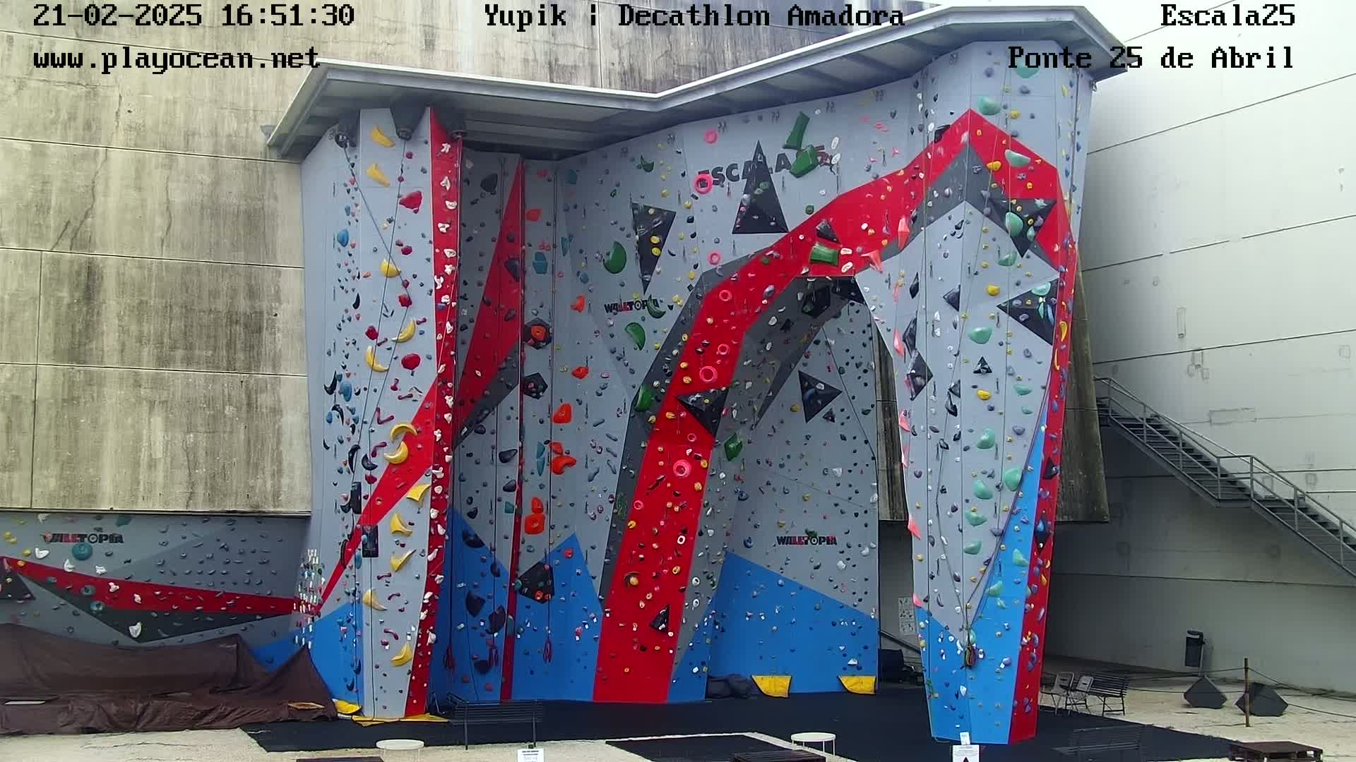 A large indoor climbing wall with various colored holds is situated against a gray building, under an overcast sky.