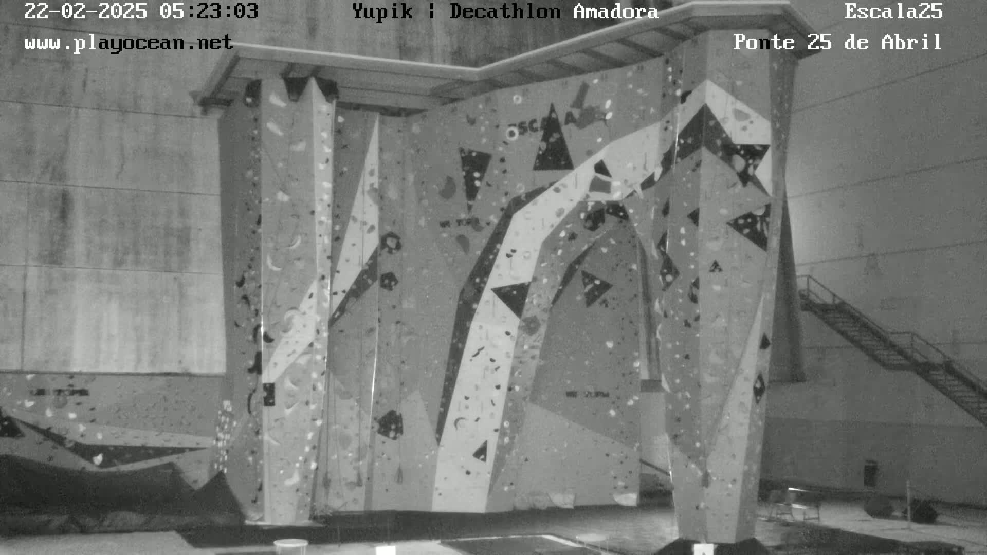 A grayscale indoor image shows a large, multi-faceted climbing wall with various colored holds.