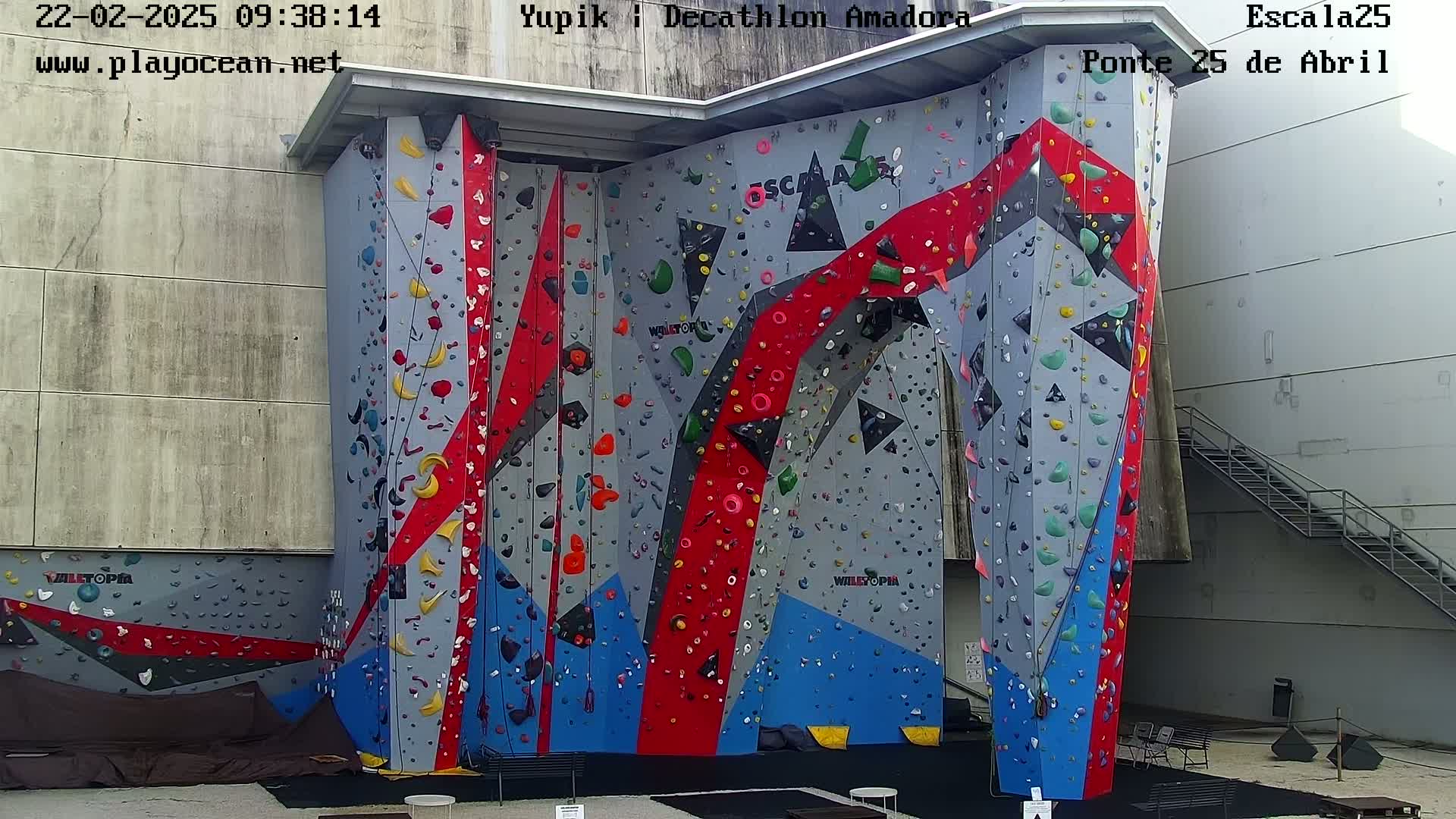 An outdoor climbing wall with various colored holds, set against a light gray building under a cloudy sky.