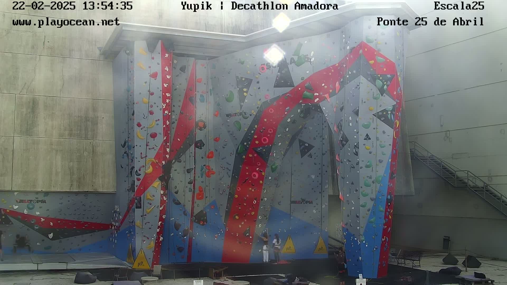 A large, multi-colored indoor climbing wall stands against a light gray building on a somewhat hazy day.