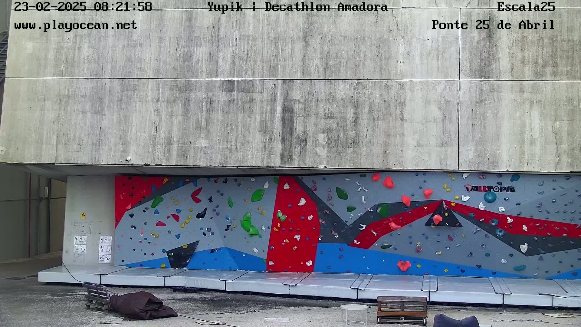 An outdoor climbing wall with multicolored holds is situated against a concrete building.