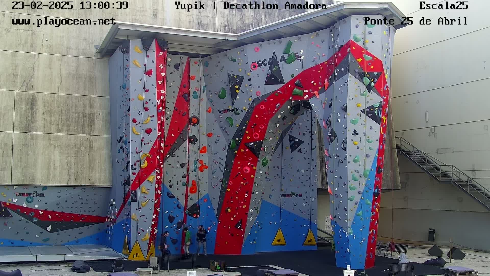 A large, colorful outdoor climbing wall with several people standing at its base.