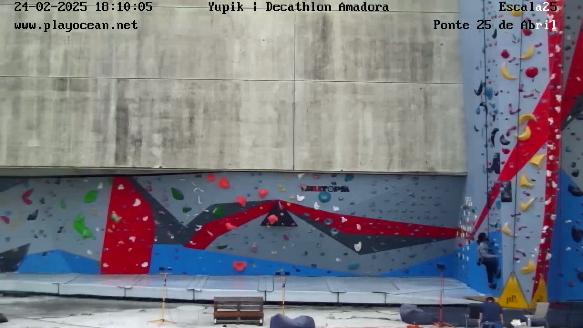 A person climbs an outdoor climbing wall with multicolored holds against a concrete building.