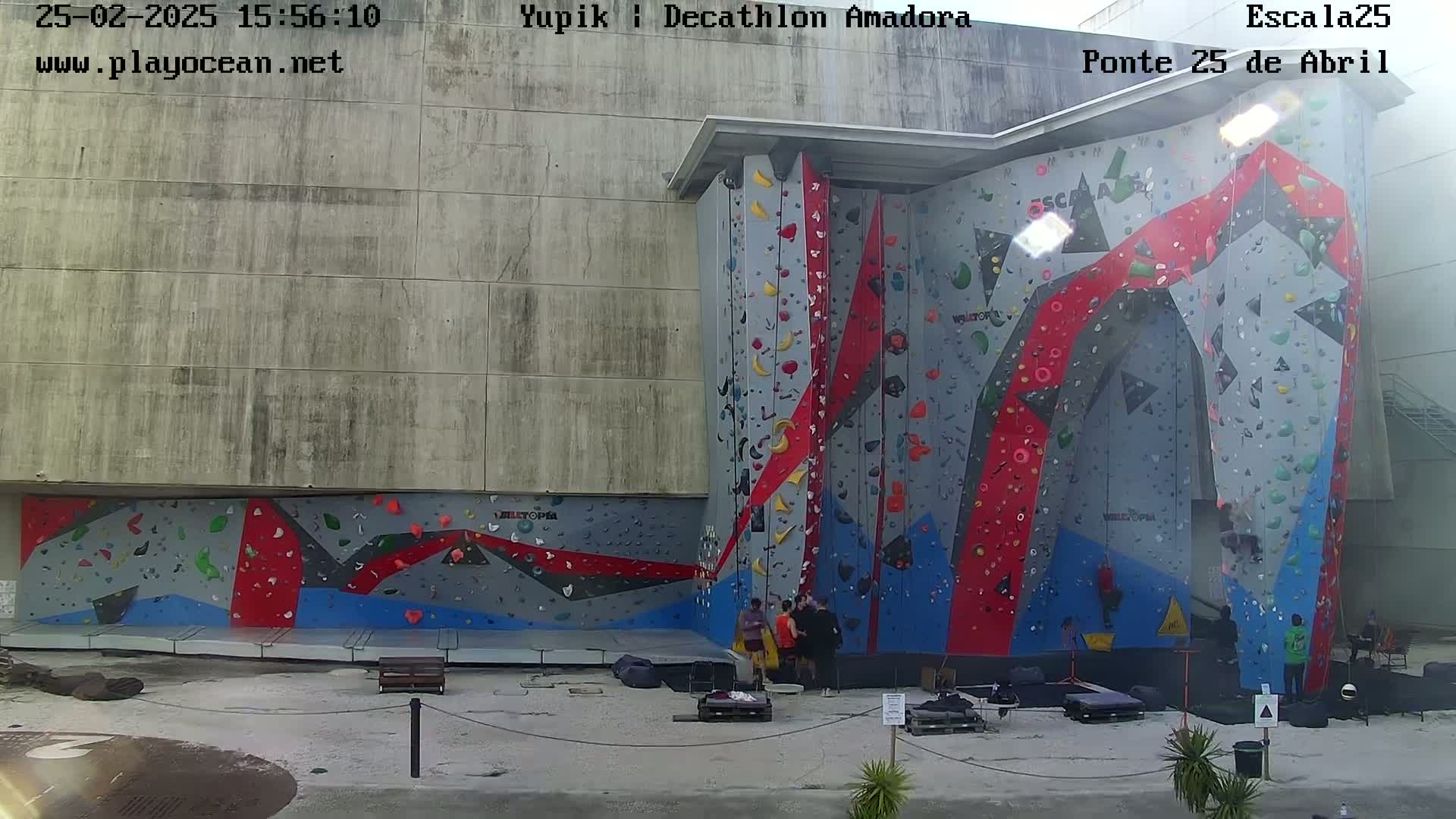 A large outdoor climbing wall with various colored sections, mostly gray, red, and blue, is surrounded by a paved area with a few people near the base.