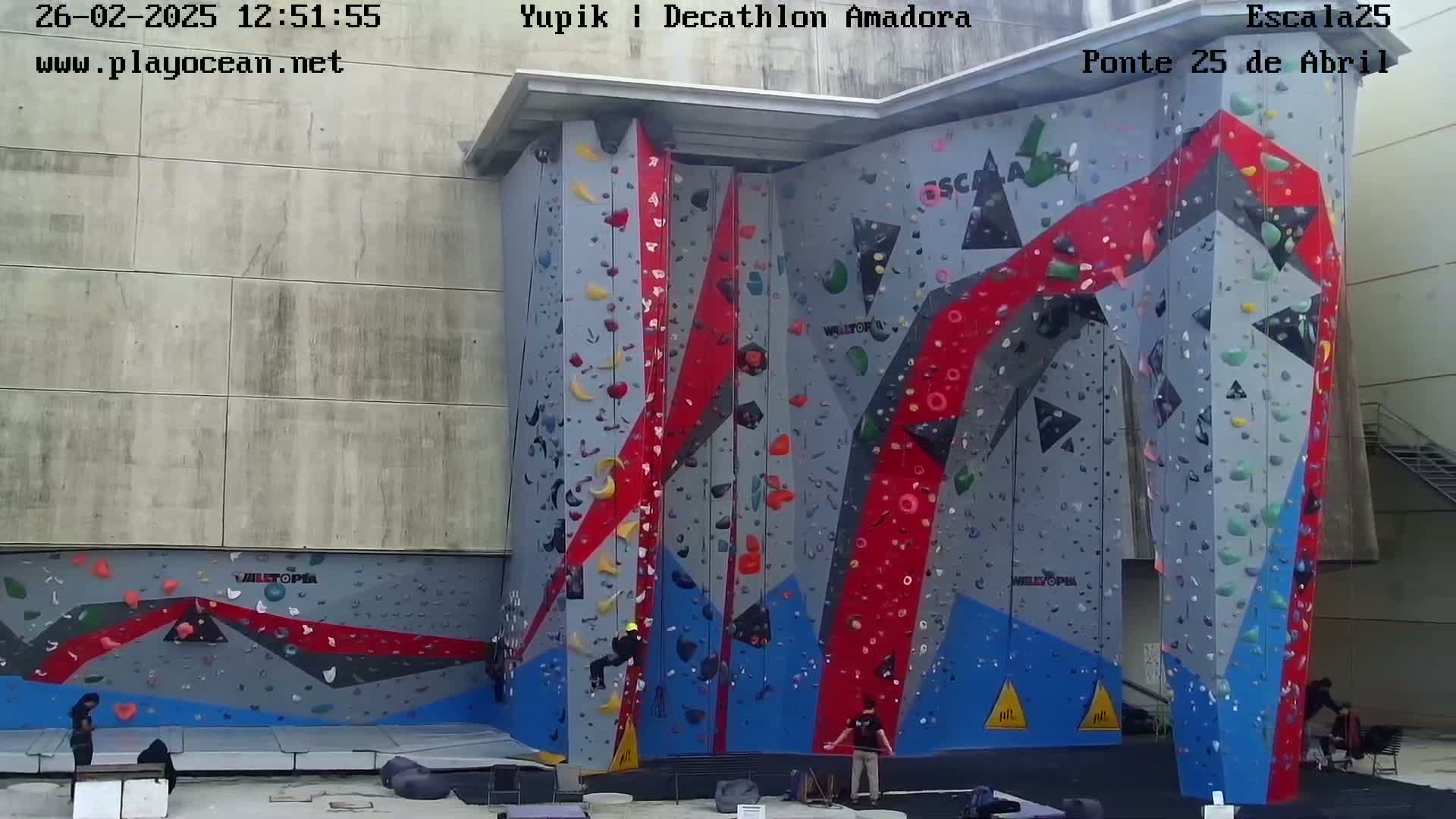 Several people climb a large, colorful outdoor climbing wall on a mostly cloudy day.