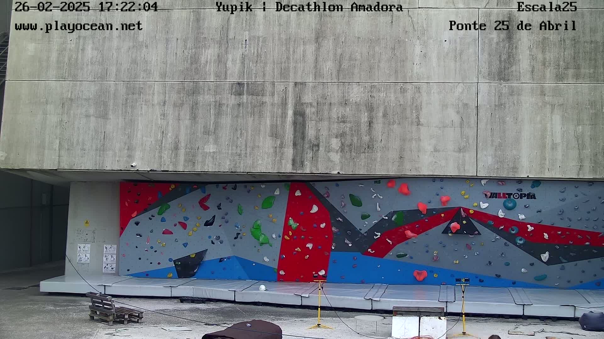 A large outdoor climbing wall with various colored holds is situated beneath a concrete overhang.