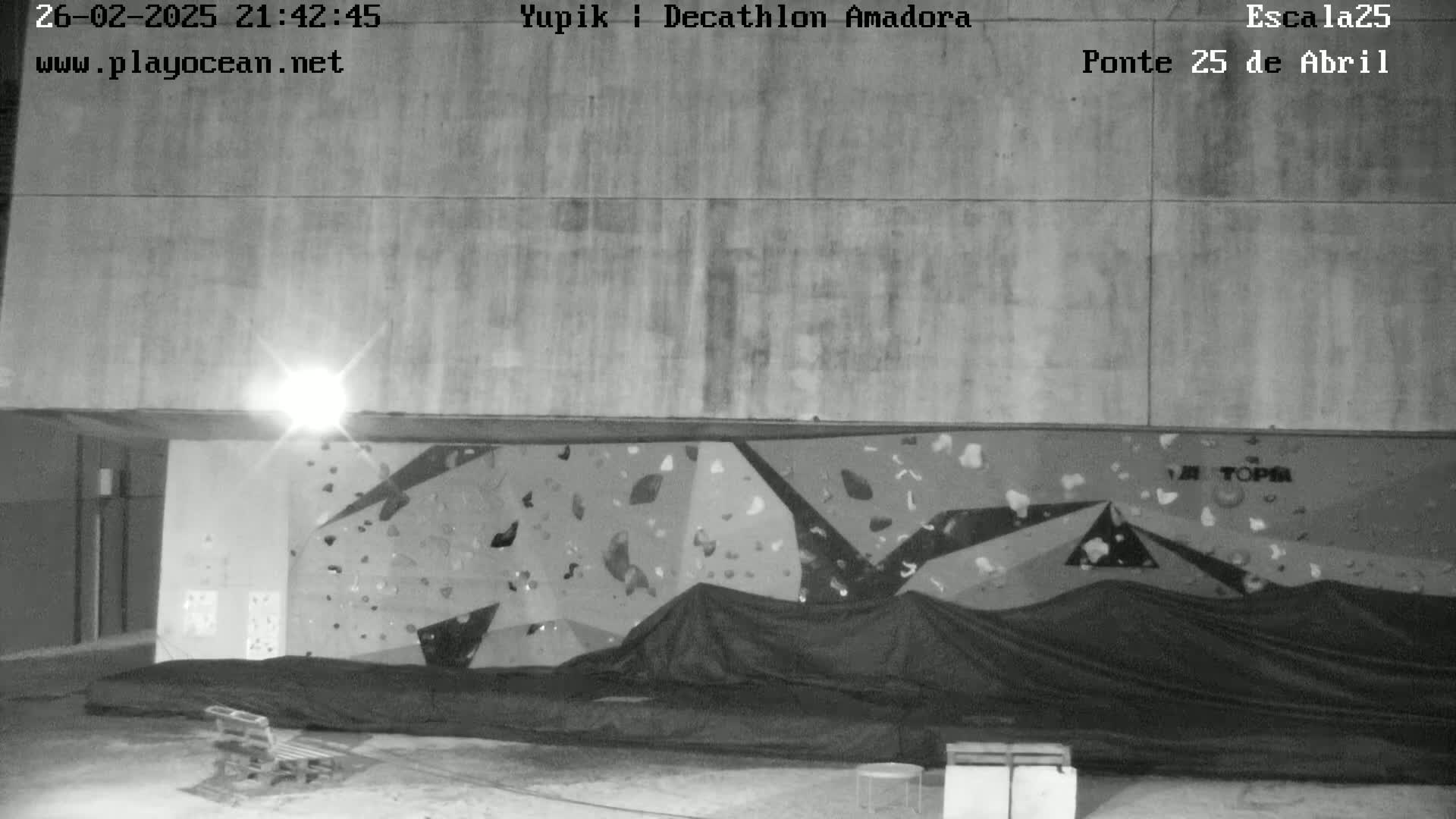 A nighttime, black-and-white image shows an outdoor climbing wall covered by a tarp, with a single bench visible in the foreground.