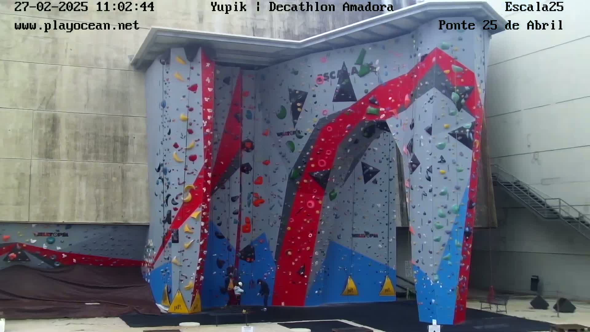 An outdoor climbing wall with red, blue, and gray sections is visible, with a few people near the base on an overcast day.