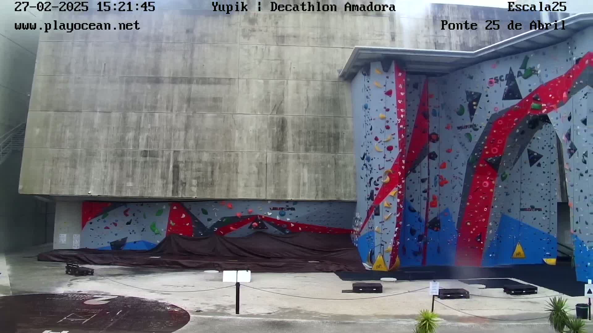 A large outdoor climbing wall with red, blue, and gray holds is partially covered by a dark tarp, and the area is mostly empty under an overcast sky.