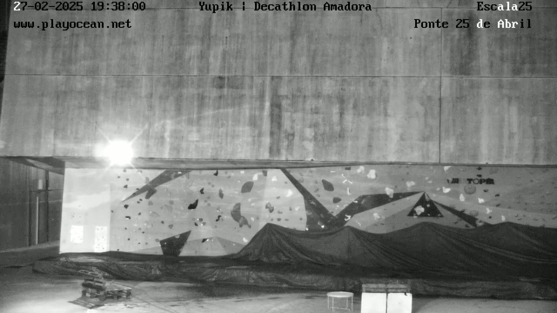A nighttime black and white image shows an outdoor climbing wall partially covered by a tarp, situated beneath a large concrete structure.