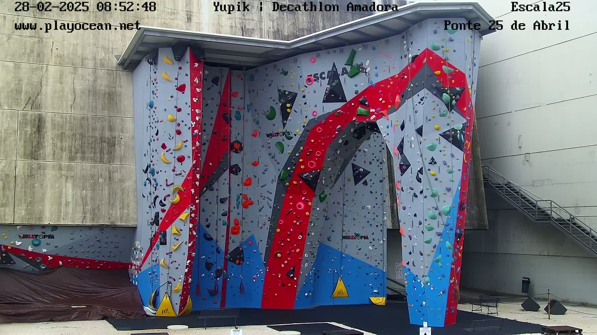 A large, colorful indoor climbing wall stands against a light gray building on a partly sunny day.