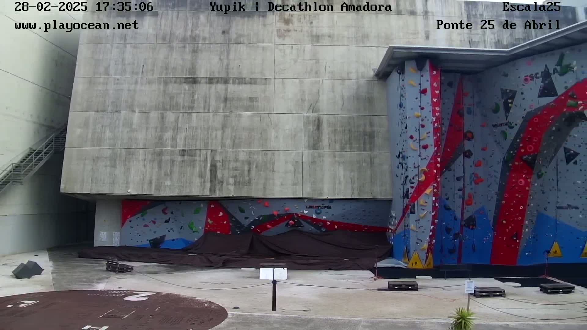 A large outdoor climbing wall with various colored holds is situated next to a concrete building on a mostly clear day.