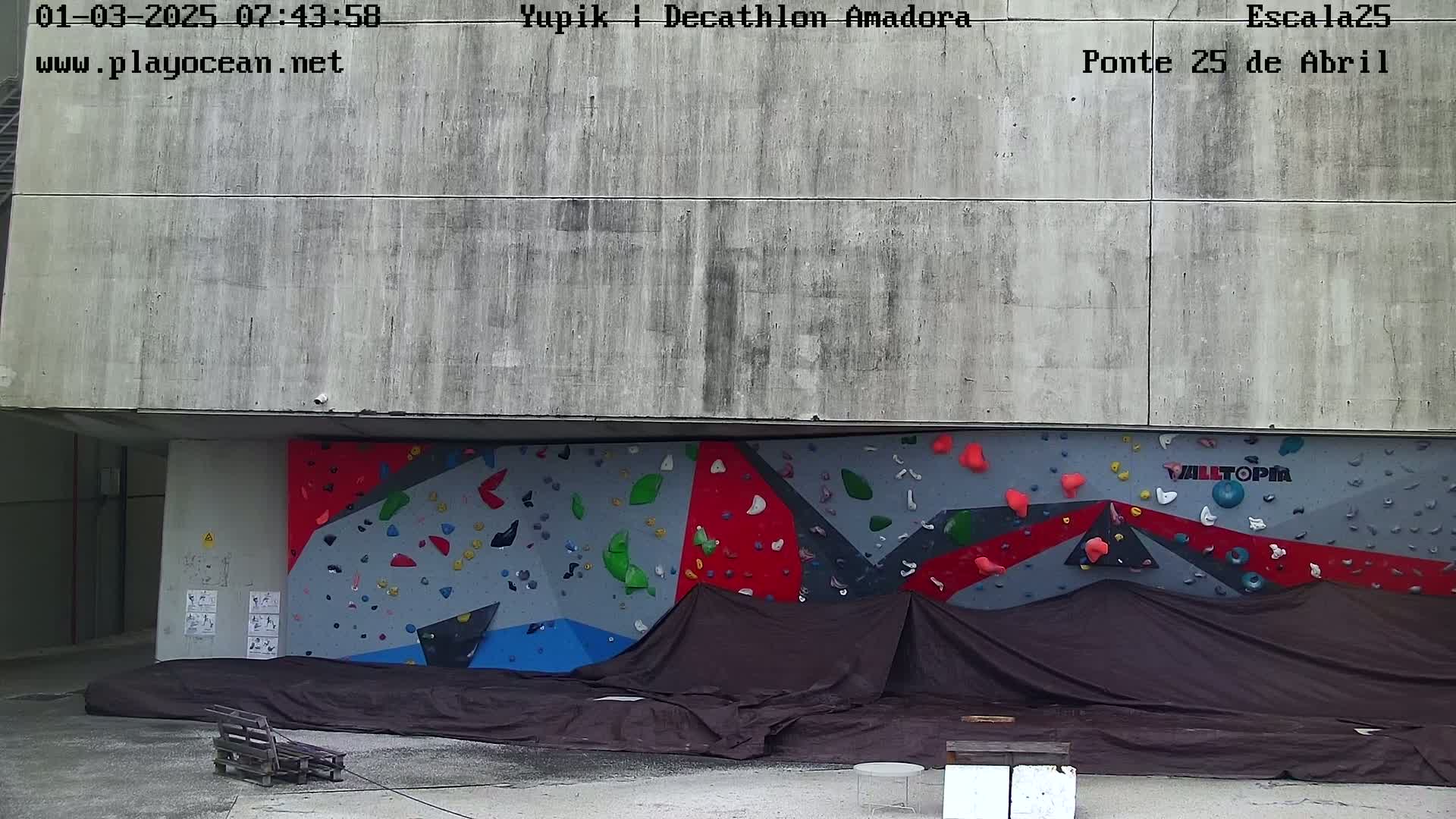 A large, colorful climbing wall is partially covered by a dark tarp, situated against a weathered concrete building outdoors.