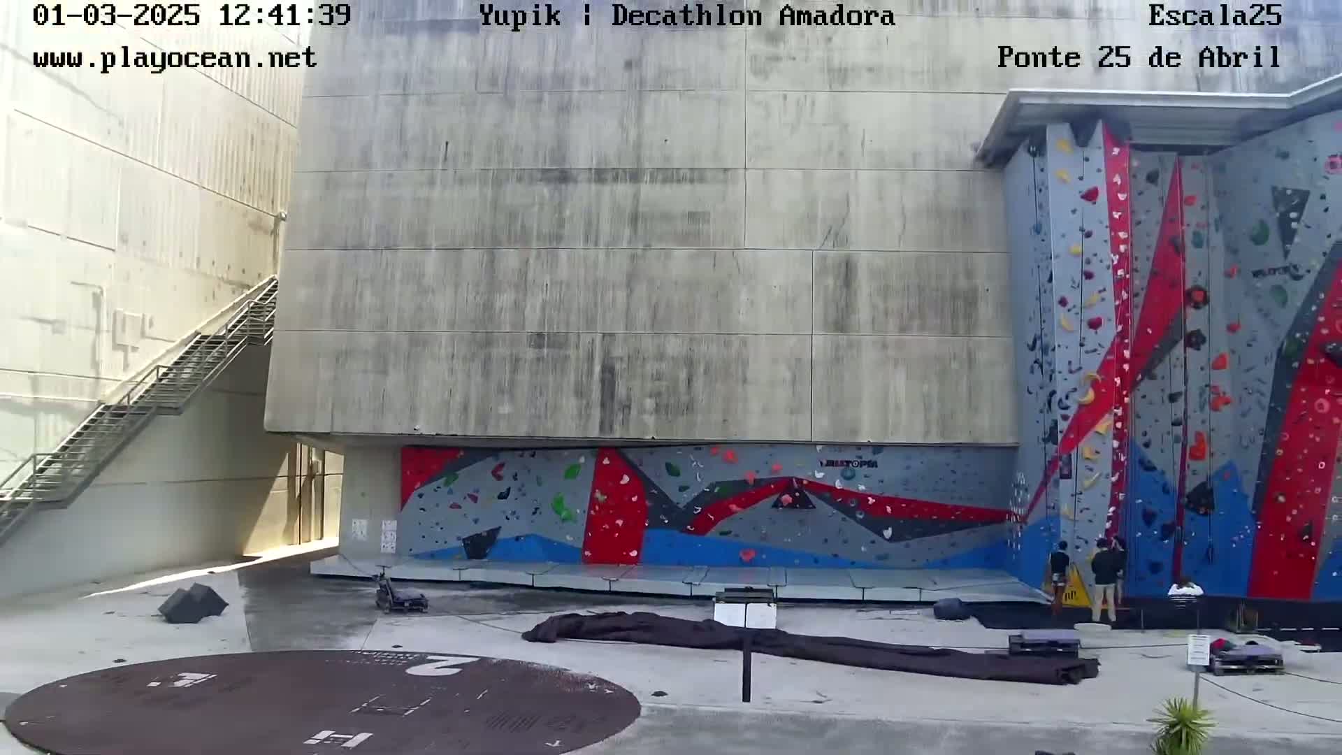 A large, outdoor climbing wall with colorful holds is situated next to a concrete building with a metal staircase, under sunny conditions.
