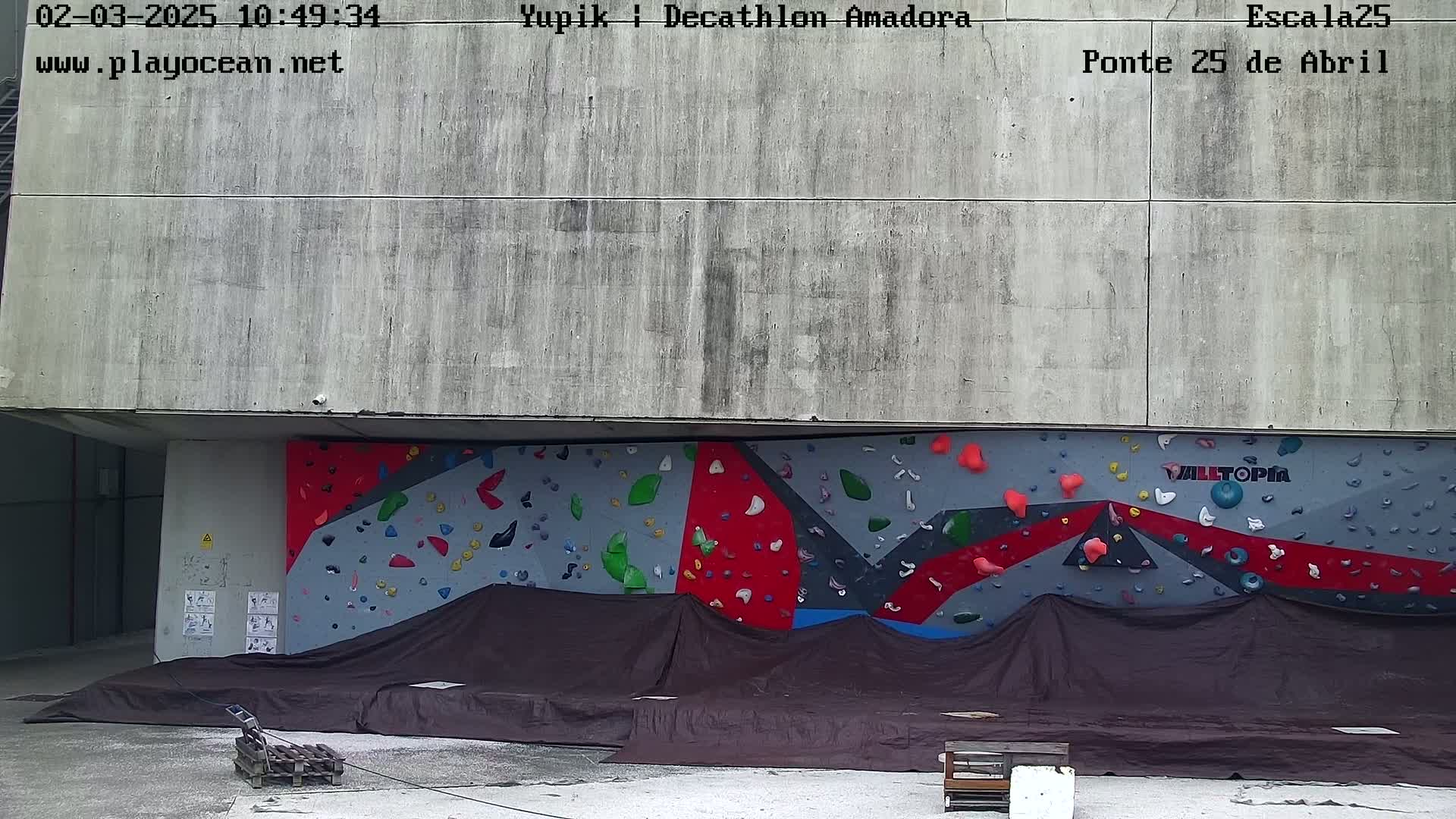 A large, multicolored climbing wall is partially covered by a dark tarp, situated against a concrete building outdoors.