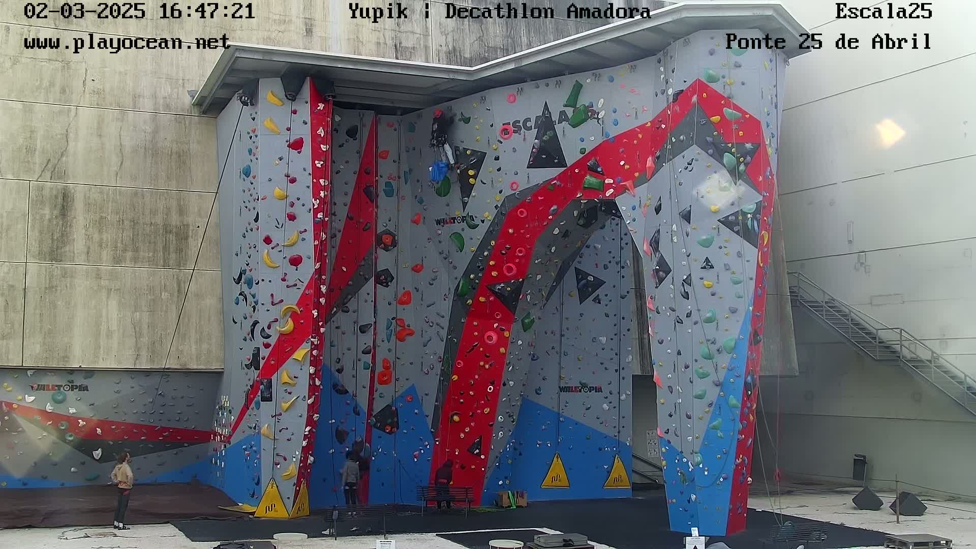A large, multi-colored indoor climbing wall stands against a light-grey building under an overcast sky.