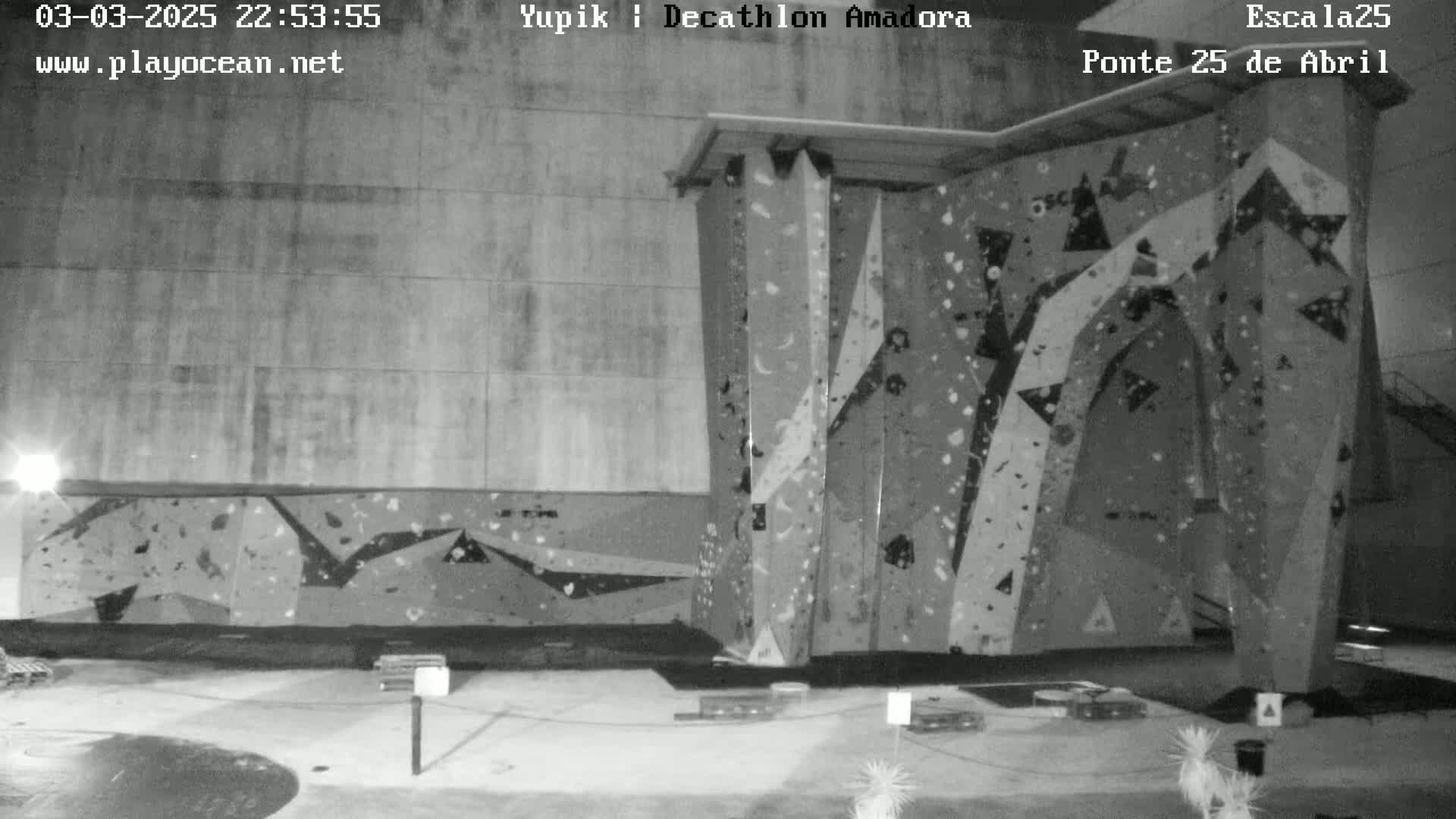 A nighttime black and white image shows an indoor climbing wall with various holds and features, under artificial lighting.