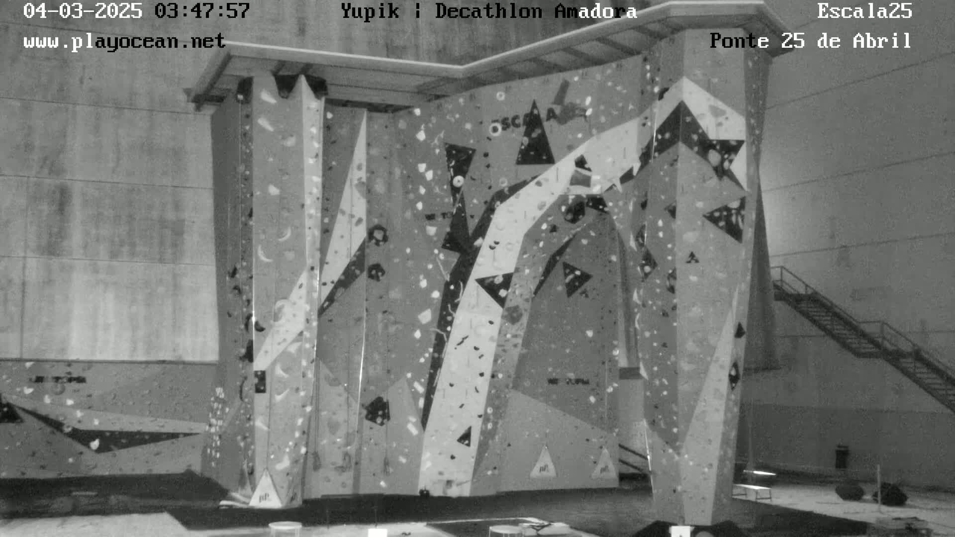 A grayscale indoor image shows a large indoor climbing wall with various holds and geometric patterns.