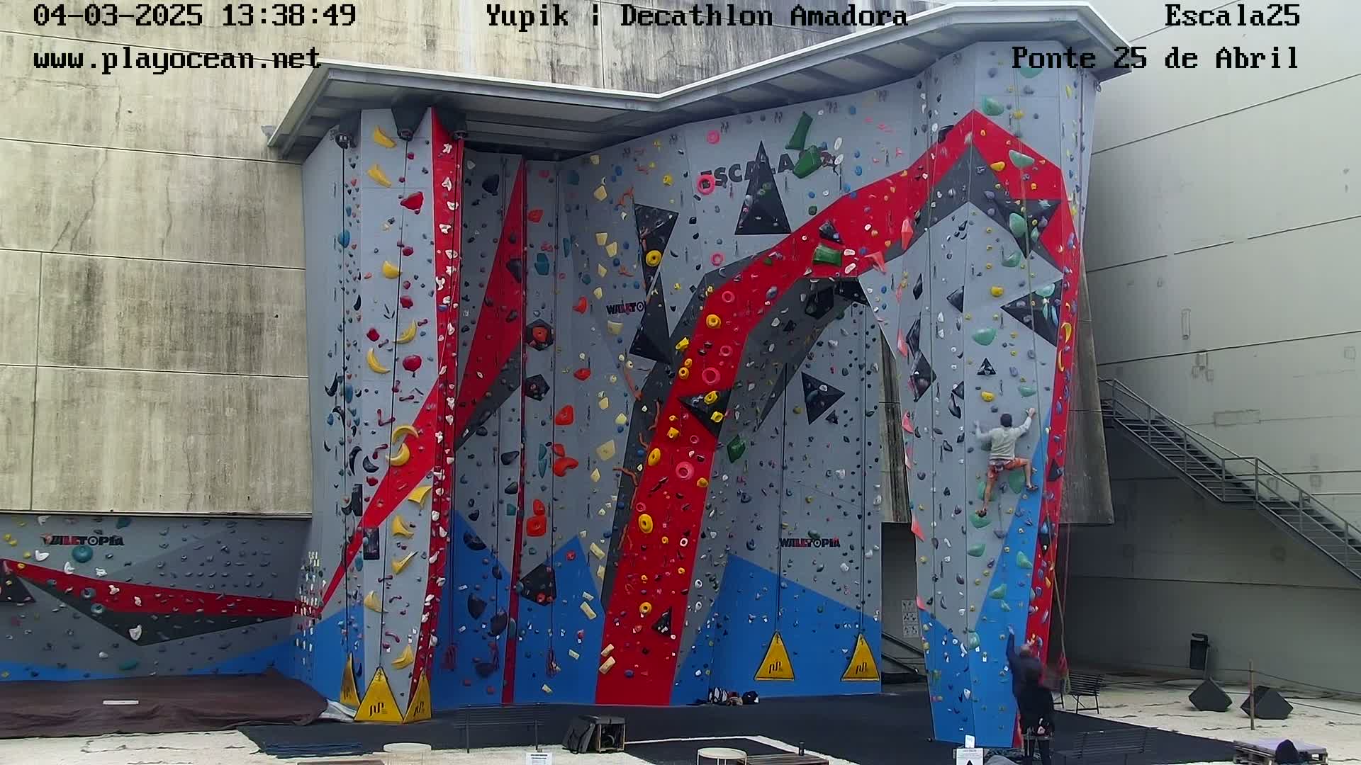 A large outdoor climbing wall with various colored holds and a person climbing.