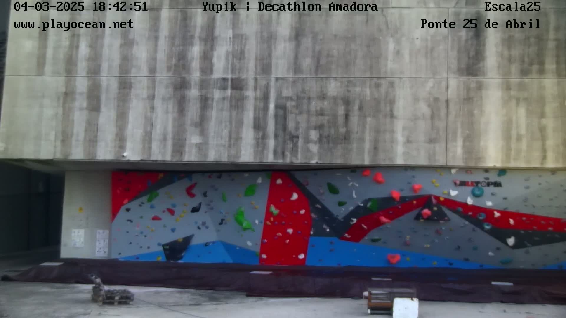 A large outdoor climbing wall with red, blue, and gray sections is situated beneath a concrete structure.