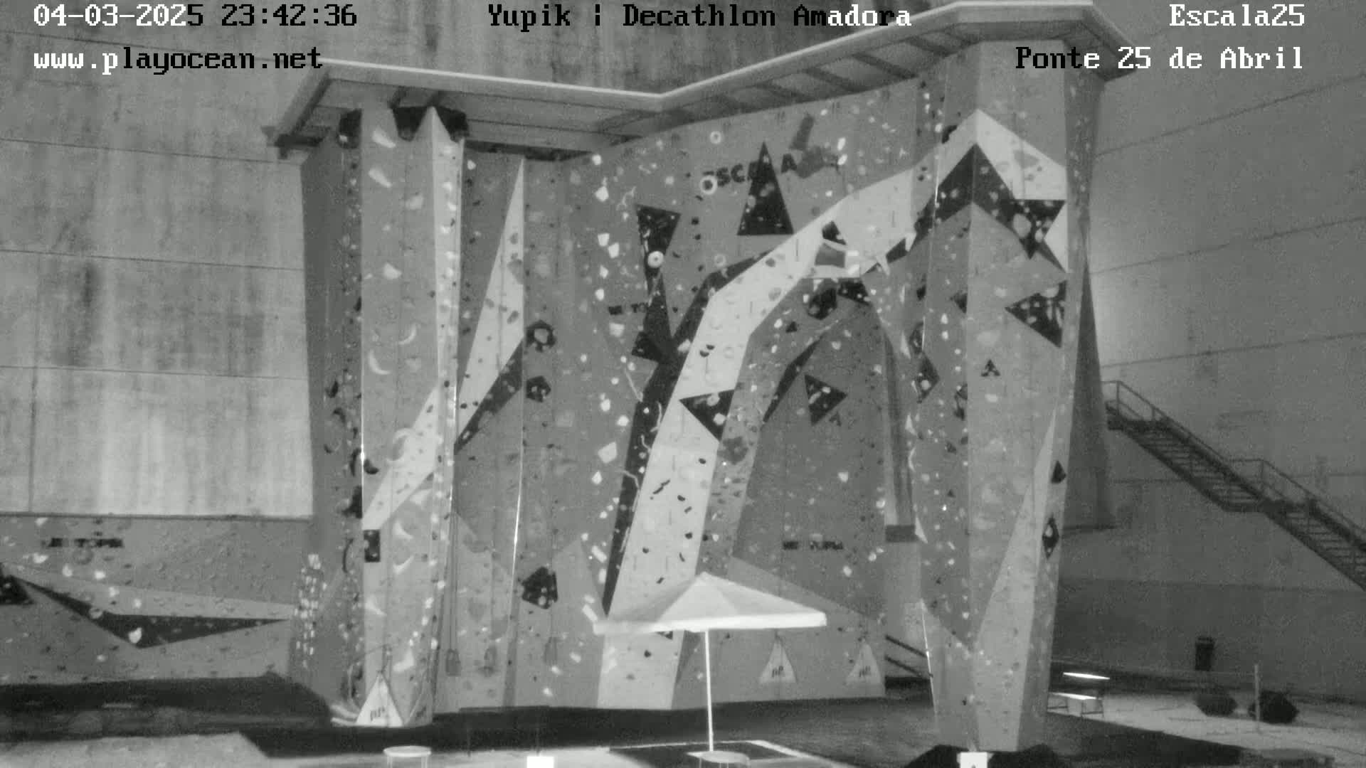 A large indoor climbing wall with various holds and a closed umbrella sits in a concrete structure.