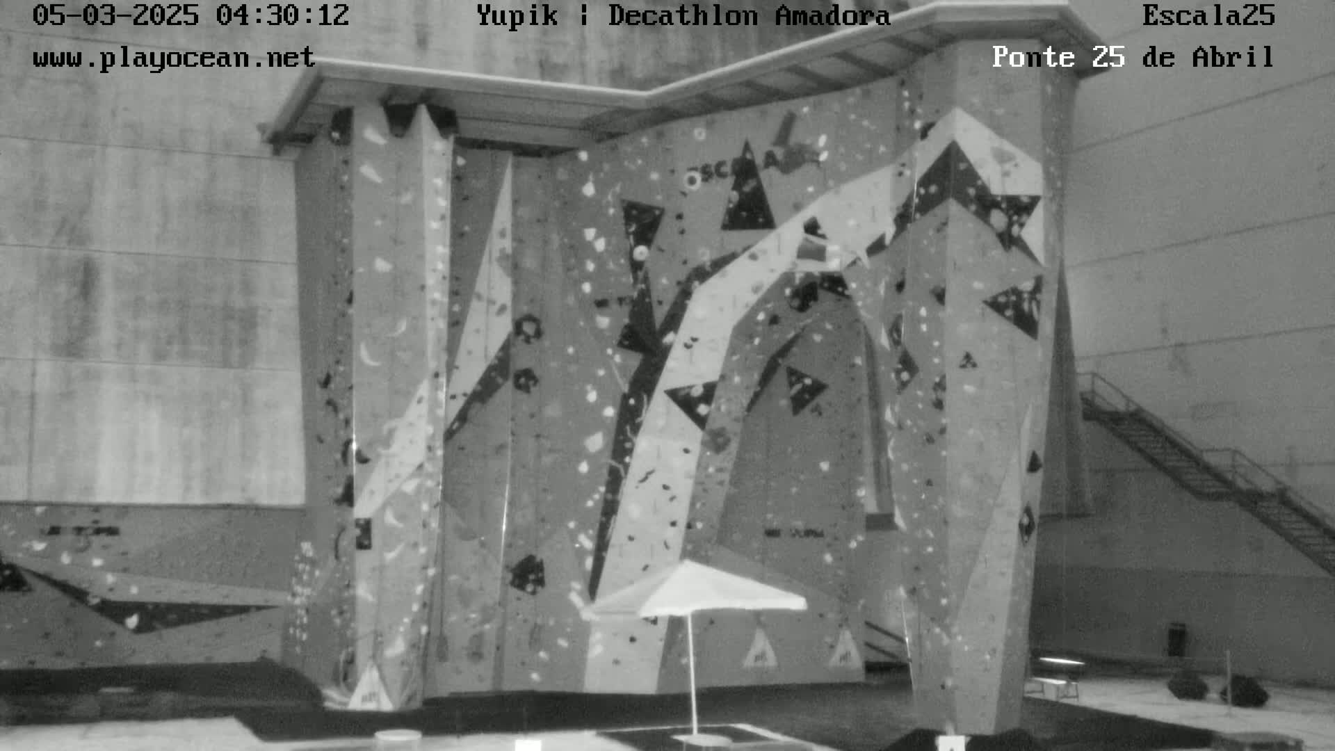 A large indoor climbing wall with various holds and a closed umbrella sits in a grey-toned room.