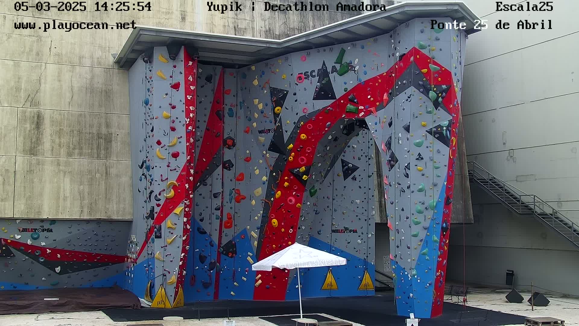 A large outdoor climbing wall with various colored holds and a white umbrella is situated next to a concrete building with a metal staircase.