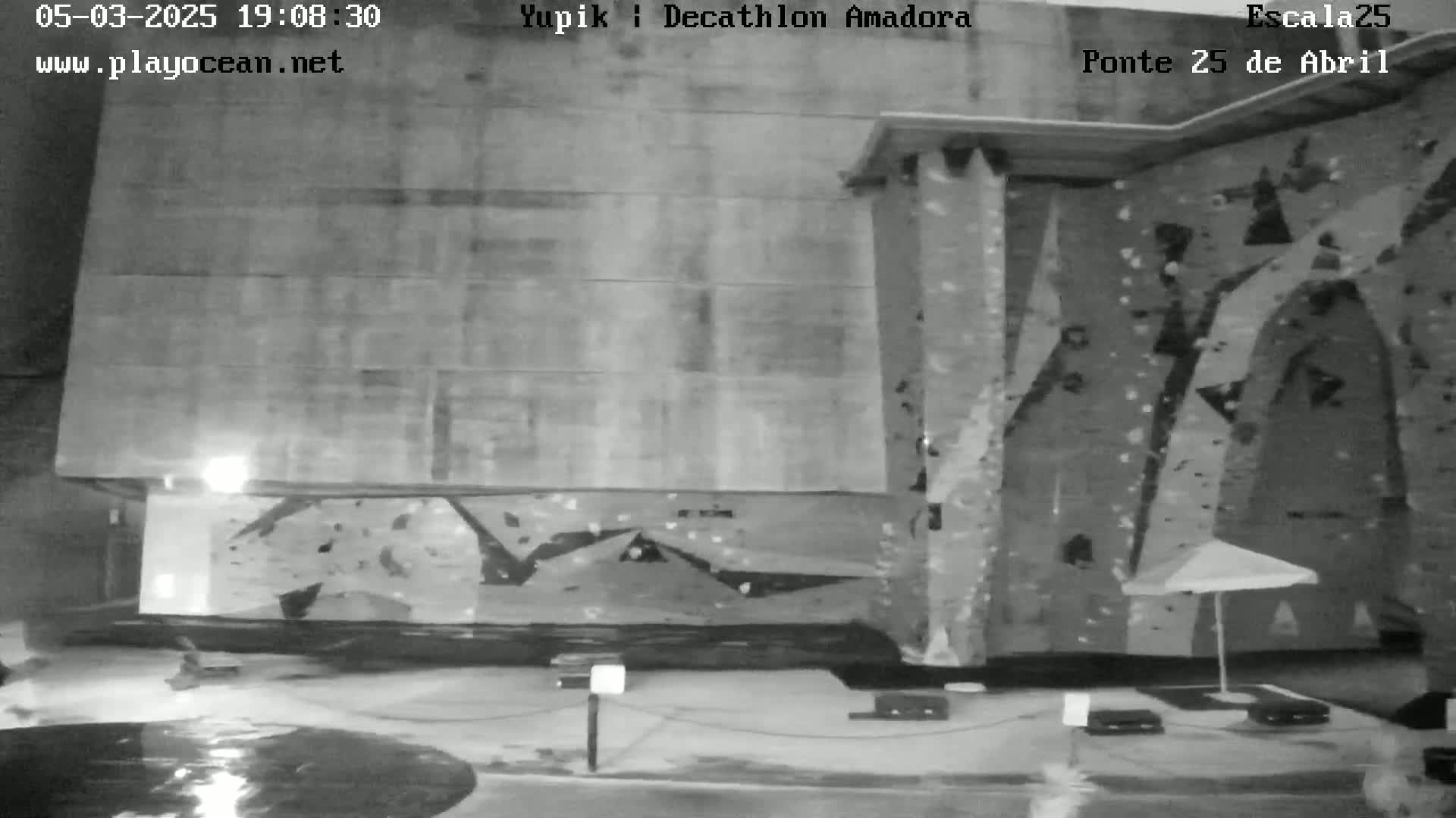 A nighttime, black-and-white image shows a large outdoor climbing wall with a closed umbrella and wet ground in front.