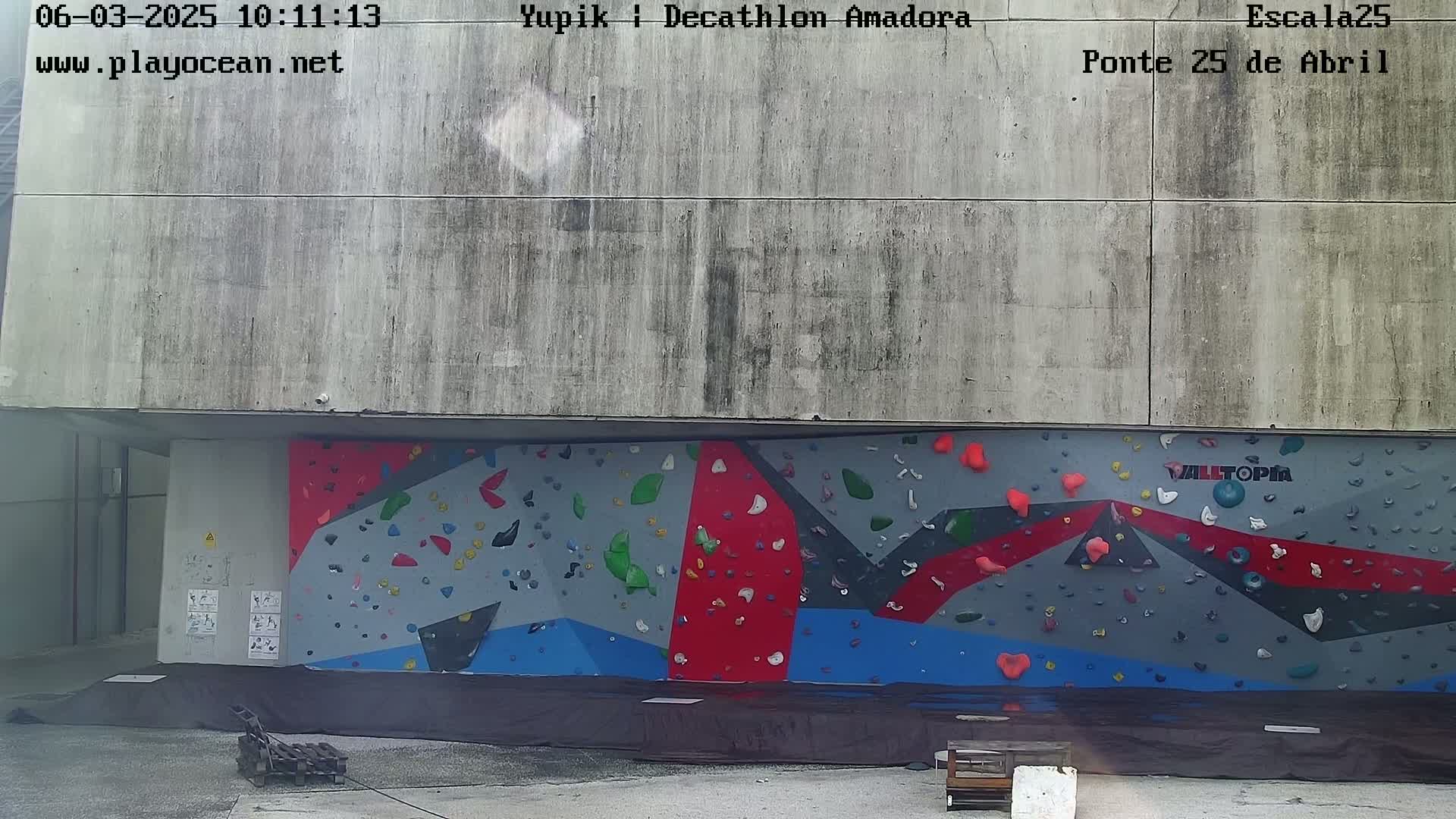 A large, outdoor climbing wall with multicolored holds is situated beneath a weathered concrete structure.