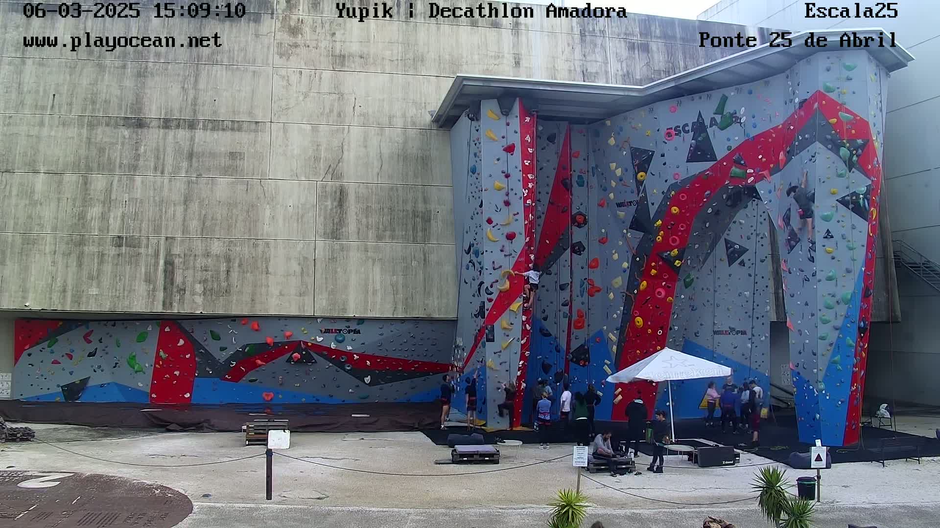 An outdoor climbing wall with several people climbing and watching, under a partly cloudy sky.