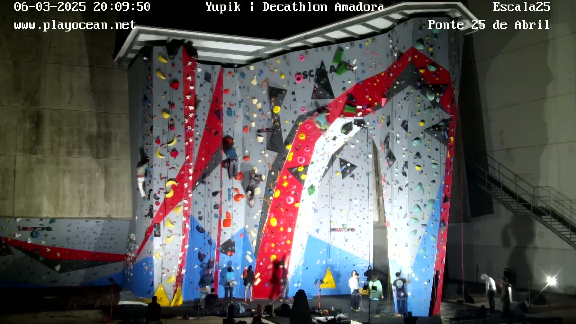 Several people are indoor rock climbing on a large, brightly colored climbing wall at night.