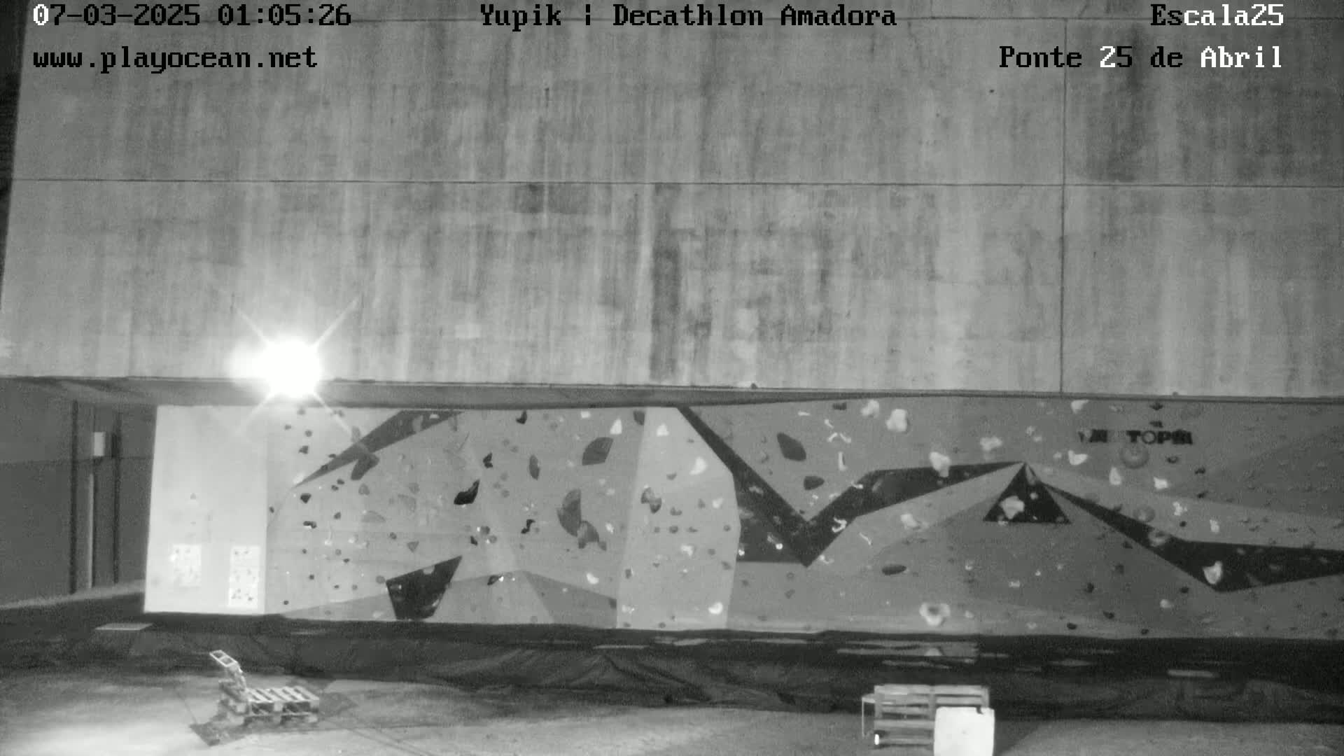 A nighttime grayscale image shows a large, outdoor climbing wall under a bright light.