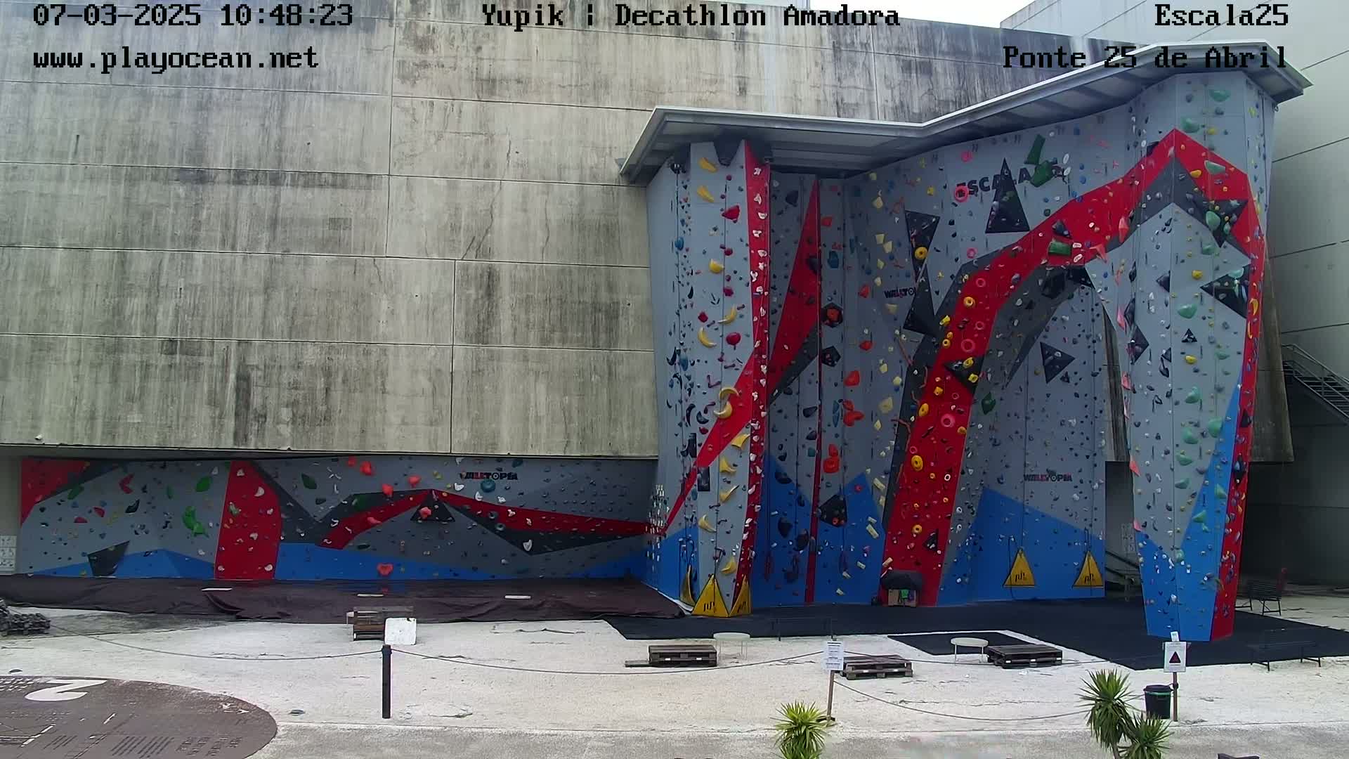 A large outdoor climbing wall with various colored holds is attached to a concrete building.
