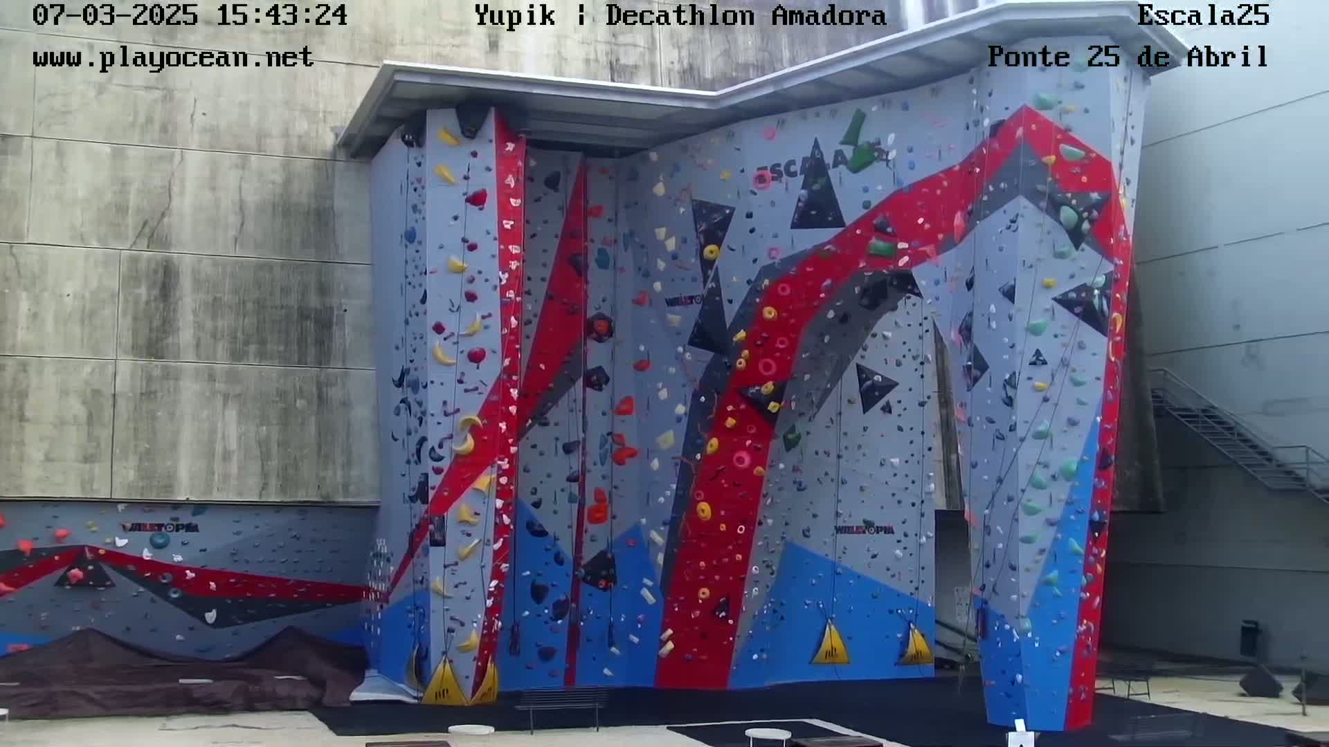 An outdoor, multi-colored climbing wall stands against a light gray building under a mostly clear sky.